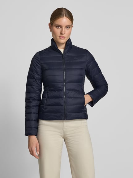 Frauenmodel trägt dunkelblaue Slim Fit Steppjacke mit Logo-Detail von Lacoste, Beigehose, glatte Oberflächen, Ganzkörperansicht von vorne, Damen Steppjacke von Lacoste.