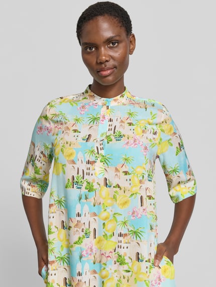 Frauenmodel trägt Knielanges Kleid aus Viskose mit 3/4-Arm von Emily Van den Bergh, gerader Schnitt, bunte Stadtmotive, Nahaufnahme Oberkörper. Rückenlänge 96 cm, Brustweite 50 cm, Ärmellänge 36 cm, reine Viskose, kurze Knopfleiste.