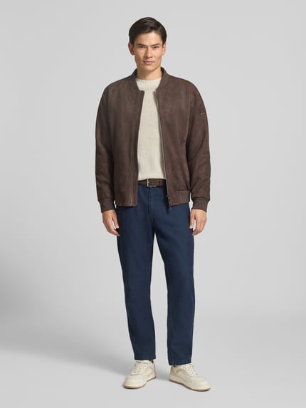 Mannenmodel draagt Slim Fit gebreide pullover uit zuiver wol met labelstitching van MCNEAL, ribboorden, ruglengte maat M 66 cm, mouwlengte maat M 69 cm, heren, neutrale kleuren, zachte textuur, donkerbruine jas, blauwe broek, crèmekleurige schoenen, volledig aanzicht van voren.