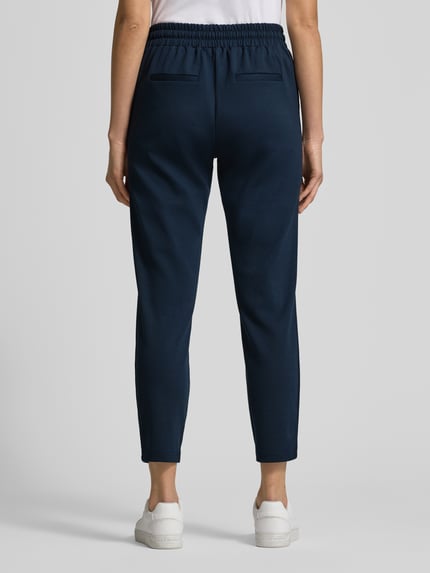 Vrouwenmodel draagt donkerblauwe sweatpants met siernaden, elastische band met tunnelkoord, model 'RIZET' van b.young, los model, materiaalmix met viscose, sierzakken achter, detail van de broek getoond, geheel lichaam achteraanzicht.
