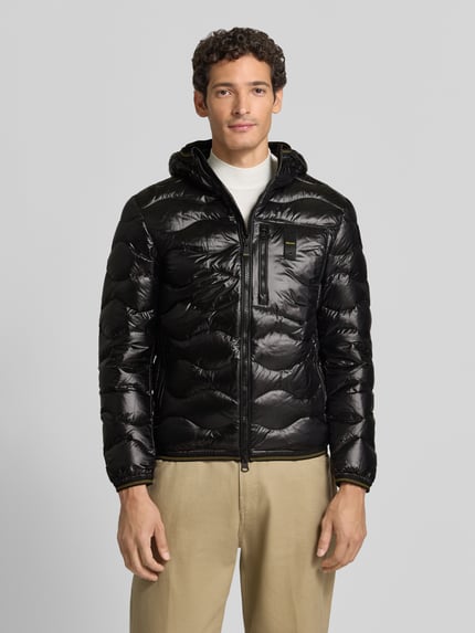 Männermodel trägt schwarze Steppjacke mit Zweiwege-Reißverschluss Modell 'WAVE' von Blauer USA, Kontrast zu beiger Hose, Ganzkörperansicht von vorne. Herren Steppjacke Slim Fit mit Kapuze, Label-Detail sichtbar, glatte glänzende Textur, Kontext: Rückenlänge 68 cm, Schulterbreite 47 cm, Brustweite 53 cm, Ärmellänge 49 cm.