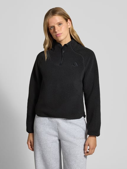 Frauenmodel trägt schwarze Regular Fit Fleecejacke mit Troyerkragen von adidas Sportswear, dezente Textur, Ganzkörperansicht von vorne.