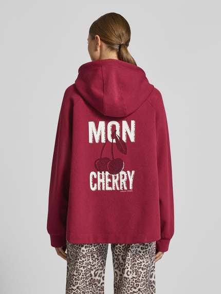 Männermodel trägt Relaxed Fit Hoodie mit Motiv-Print von Marc Cain, elastyczna mieszanka bawełny, wzór statement, kaptur ze sznurkiem, elastyczne mankiety, widok całej sylwetki od tyłu.