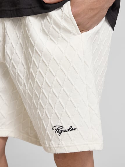 Männermodel trägt Shorts mit Logo-Stitching Modell 'Jovren' von Pegador im weißen Material-Mix mit Rautenstruktur, Nahaufnahme Oberschenkel, Herren Shorts, Loose-Fit, Elastischer Bund, Französische Eingrifftaschen, Kontext: Bundweite bei Größe M 38 cm, Innenbeinlänge 24 cm.