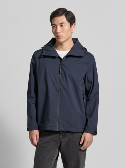 Męskimodel nosi kurtkę z kapturem i kieszeniami zapinanymi na zamek błyskawiczny GOFRANCK Jacke mit Kapuze und Reißverschlusstaschen Modell 'CYCLONE', wykonana z bawełny, w prostym kroju, kolor granatowy, detal z logo, regulowane patki przy mankietach na rzep, całkowita długość tyłu w rozmiarze M: 71 cm, szerokość ramion w rozmiarze M: 48 cm, szerokość klatki piersiowej w rozmiarze M: 49 cm, długość rękawów w rozmiarze M: 65 cm, zdjęcie w całości z przodu