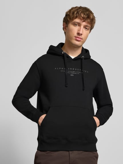 Männermodel trägt Relaxed Fit Hoodie aus Baumwoll-Mix mit Label-Detail von Alpha Industries, Kapuze, Känguru-Tasche, Nahaufnahme Oberkörper.