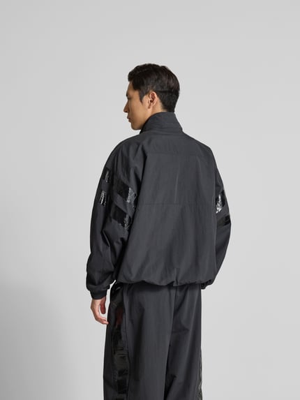 Männermodel trägt Trainingsjacke mit Stehkragen von VETEMENTS in Schwarz mit glänzenden Streifen auf den Ärmeln, Ganzkörperansicht Rückansicht, Relaxed Fit, Zweiwege-Reißverschluss, elastische Abschlüsse, Eingrifftaschen, Rückenlänge 74 cm, Schulterbreite 50 cm, Ärmellänge 73 cm.