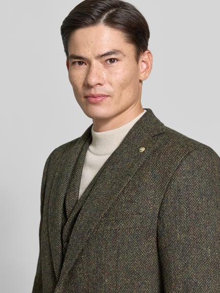 Męski model nosi Slim Fit 2-Knopf-Sakko mit Pattentaschen Modell 'Theo' od Carl Gross z czystej żywej wełny, kołnierz z połami, kieszenie z patką, kolor oliwkowy z brązowym wzorem, tekstura tkaniny wyraźnie widoczna, detaliczny widok górnej części sylwetki, Nahaufnahme Oberkörper.