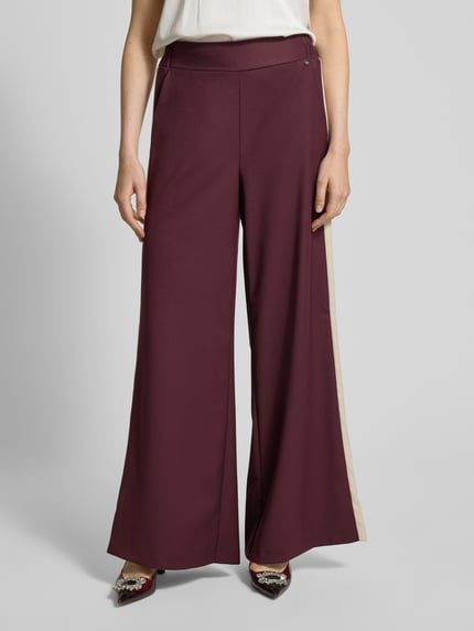 Kobieta nosi Stoffhose mit weitem Bein und elastischem Bund od Rinascimento, bordowy materiał, kieszenie, detal z logo, widok z przodu, zbliżenie na nogi.