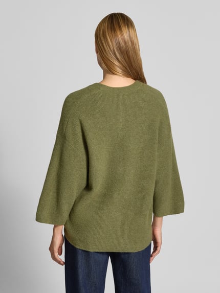 Frauenmodel trägt Oversized Cardigan aus Baumwoll-Anteil Modell 'DAMSVILLE' von American Vintage in grünlicher Strick-Optik, Ganzkörperansicht Rückansicht. Elastischer Material-Mix, Oversized Fit, Knopfleiste, V-Ausschnitt, Rückenlänge 63 cm, Ärmellänge 35 cm.