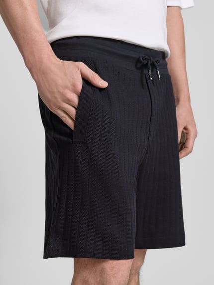 Mężczyzna nosi Regular Fit Shorts mit Tunnelzug Modell 'JUANITO' od JOOP! Collection, czarna tkanina bawełniana, szerokość w rozmiarze M 43 cm, długość nogawki 17 cm, tekstura prążkowana, zbliżenie na górną część ciała
