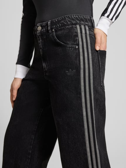 Frauenmodel trägt Loose Fit Jeans aus reiner Baumwolle von adidas Originals in schwarzem Denim mit Kontraststreifen, Nahaufnahme Hüfte.