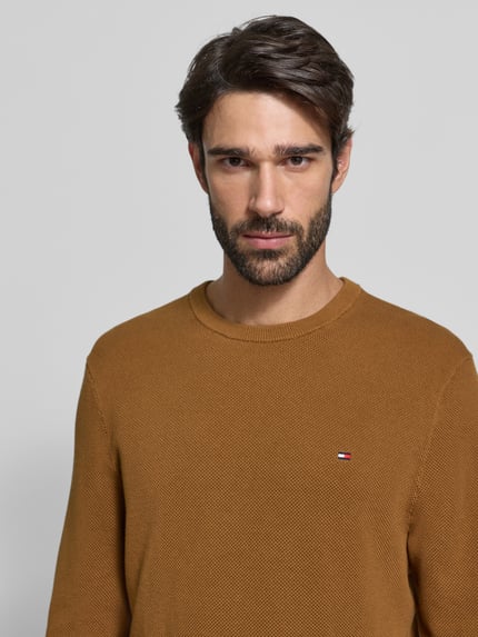 Männermodel trägt Regular Fit Strickpullover aus reiner Baumwolle von Tommy Hilfiger in Braun, feine Struktur, Logo-Detail, Nahaufnahme Oberkörper.