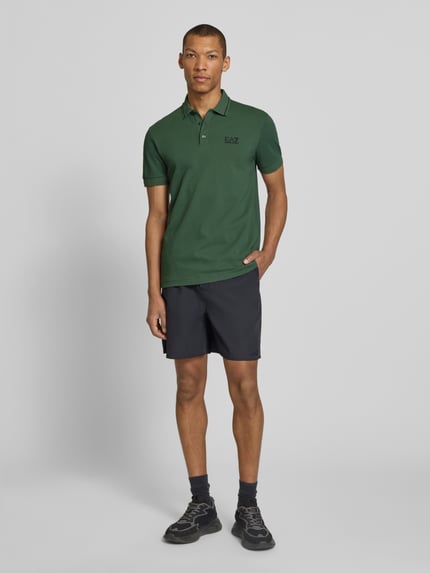 Männermodel trägt Regular Fit Poloshirt mit Logo-Print von EA7 Emporio Armani in Dunkelgrün, elastischer Baumwoll-Mix, Ganzkörperansicht von vorne.