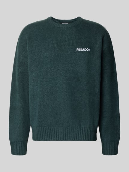 Produktfoto, Oversized Strickpullover Modell 'Luka' von PEGADOR in Dunkelgrün mit Rundhalsausschnitt, auffälligem Label-Stitching, grober Strickstruktur, Material-Mix mit Stretch-Anteil