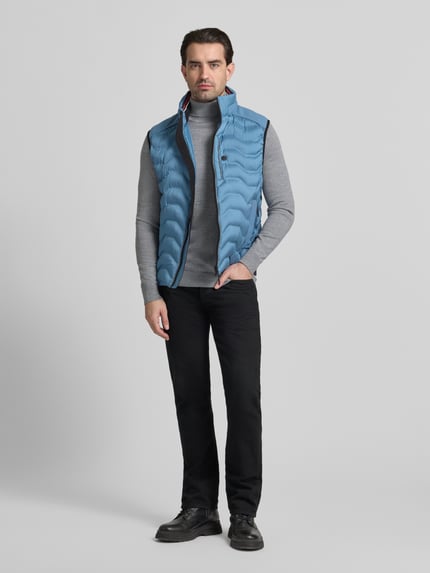 Männermodel trägt Regular Fit Steppweste mit Kapuze im Kragen von Tom Tailor in Hellblau, kombiniert mit grauem Rollkragenpullover, Ganzkörperansicht von vorne. Unifarbene Optik, reflektierende Details, Label Patch, Reißverschluss, seitliche Taschen sowie Innentasche sichtbar.