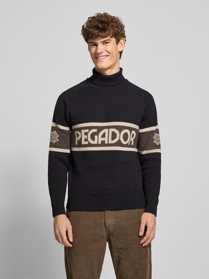 Männermodel draagt Strickpullover mit Rollkragen van Pegador in zwart met beige labelprint, katoenmix, regular fit, geribde boorden, vooraanzicht bovenlichaam.