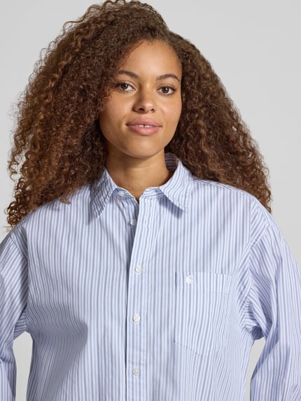 Vrouwenmodel draagt Relaxed Fit blouse met labelstitching Model 'Keene' van Carhartt Work In Progress, puur katoen, overhemdkraag, knoopsluiting, borstzak, lichtblauw-wit gestreept, textuur glad, Nahaufnahme Oberkörper. Ruglengte bij maat S: 78 cm, mouwlengte bij maat S: 56 cm.