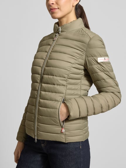 Modelka kobieca nosi Kurtka pikowana z zamkiem błyskawicznym dwustronnym model 'JUDY' od Frieda & Freddies w kolorze oliwkowym, krój slim fit, stójka, kieszenie zapinane na zamek błyskawiczny, długość tyłu w rozmiarze 36: 57 cm, szerokość ramion w rozmiarze 36: 40 cm, szerokość klatki piersiowej w rozmiarze 36: 48 cm, długość rękawów w rozmiarze 36: 64 cm, całkowity widok z przodu.