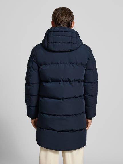 Männermodel trägt Mantel mit Kapuze und Label-Patch von NAUTGÅRD in Dunkelblau, Steppung, Rückenansicht, gerader Schnitt, Ganzkörperansicht Rückansicht.