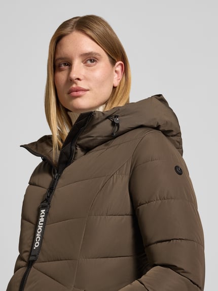 Vrouwenmodel draagt Regular fit gewatteerde mantel met capuchon Model 'SATZE' van khujo, doorgestikte naden, steekzakken, tweewegritssluiting, kleur olijfgroen, detailopname bovenlichaam. Ruglengte maat S: 98 cm, schouderbreedte 42 cm, borstwijdte 47 cm, mouwlengte 60 cm.