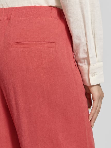 Frauenmodel trägt Stoffhose aus Viskose-Leinen-Mix mit elastischem Bund von Jake*s Casual in Koralle, Nahaufnahme Rückansicht. Relaxed-Fit mit Tunnelzug, breite Bundweite von 36 cm bei Größe 36, glatte Textur. Damen Stoffhose aus Viskose-Leinen-Mix mit Innenbeinlänge 100 cm.