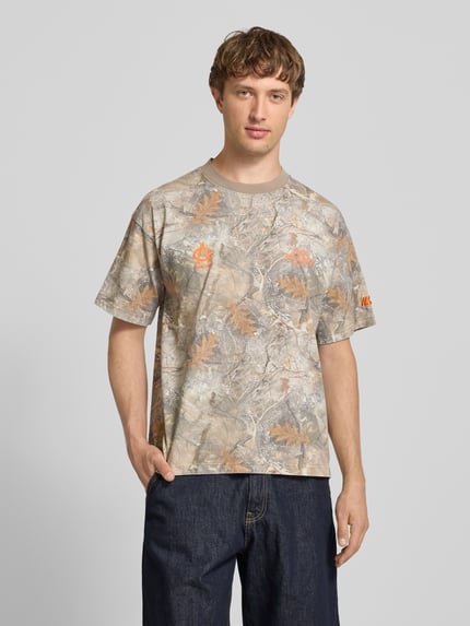 Męski model nosi Relaxed Fit T-Shirt mit Logo-Print od ANOTHER COTTON LAB, bawełna, kwiatowy nadruk na całej powierzchni, krój relaxed fit, krótki rękaw, długość tyłu 68 cm w rozmiarze M, widok w pełnej sylwetce z przodu.