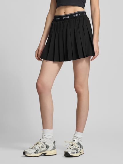 Frauenmodel trägt Faltenrock mit elastischem Bund Modell 'SELENE' von Guess Activewear, schwarzer Minirock in A-Linie, Material-Mix mit Stretch-Anteil, Bundweite 35 cm, Gesamtlänge 36 cm, Ganzkörperansicht von vorne.