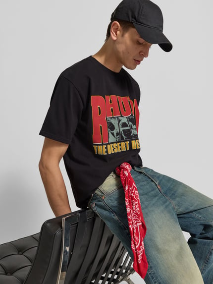 Männermodel trägt T-Shirt mit Label-Print von RHUDE in Schwarz, lockerer Schnitt, farbiger Aufdruck, reine Baumwolle, Nahaufnahme Oberkörper.