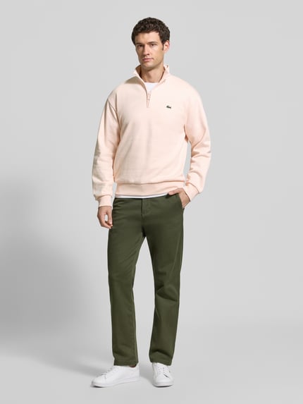 Męski model nosi Regular Fit Troyer aus Baumwoll-Mix von Lacoste w jasnoróżowym kolorze, oliwkowe spodnie, białe buty, Ganzkörperansicht von vorne. Herren Troyer von Lacoste