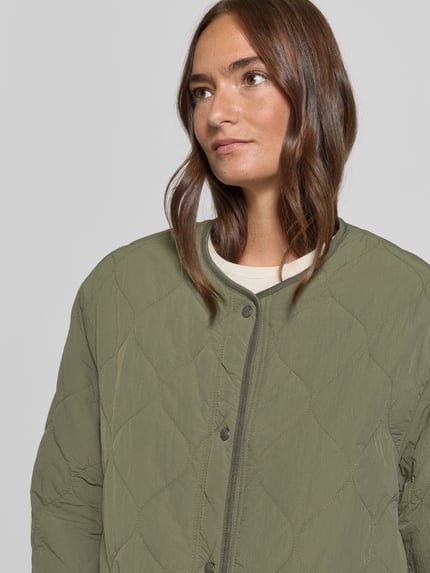Kobiece model nosi Wendejacke mit Rundhalsausschnitt od camel active w kolorze oliwkowym, wyraźna tekstura, krój regular fit, okrągły dekolt, listwa guzikowa, nakładane kieszenie, długość tyłu w rozmiarze 36: 68 cm, szerokość ramion w rozmiarze 36: 51 cm, szerokość klatki piersiowej w rozmiarze 36: 59 cm, długość rękawów w rozmiarze 36: 59 cm, zbliżenie na górną część ciała.