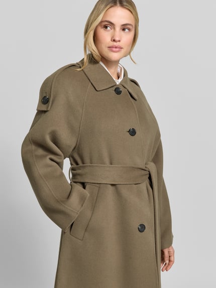 Frauenmodel trägt Mantel mit Bindegürtel Modell 'AQUALUNG' von IRO in unifarbener Optik, strukturierte Oberfläche, Oversized Passform, Teilungsnähte, Knöpfe, Umlegekragen, Schulterklappen, Langarm mit ausgestelltem Abschluss, Gehschlitz, Longline-Länge, Webware, Nahaufnahme Oberkörper.