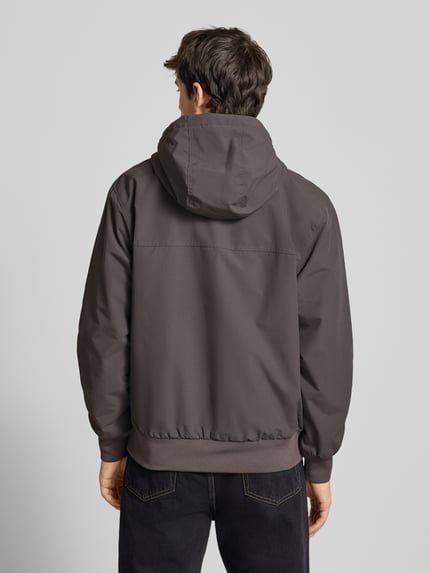 Männermodel trägt Jacke mit Kapuze von Carhartt Work In Progress in Grautönen, Regular Fit, Reißverschlusstaschen, Rückansicht, Ganzkörperansicht.