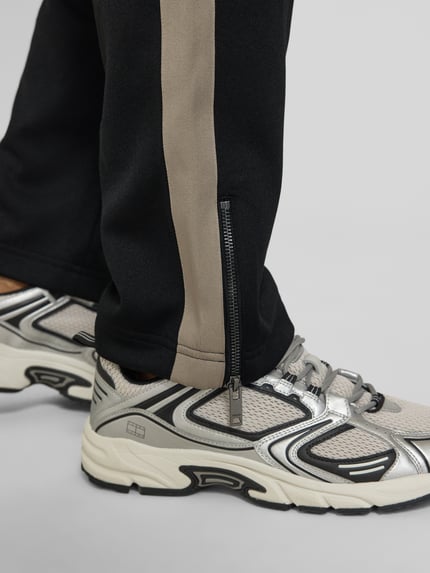 Männermodel trägt Regular Fit Jogpants mit Baumwoll-Anteil Modell 'DOLOS' von HUGO, schwarz mit beigem Kontraststreifen, Sweat-Textur, Reißverschluss am Bein, Label Print, Nahaufnahme Unterschenkel