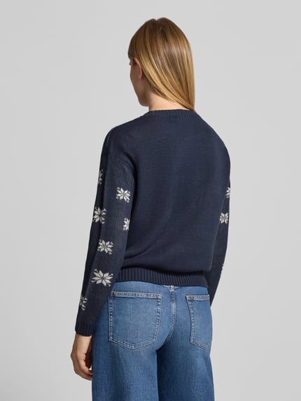 Frauenmodel trägt Pullover mit Pailletten-Schriftzug von VERO MODA in Strickoptik, mehrfarbig mit Sternenmuster, Ganzkörperansicht Rückansicht.