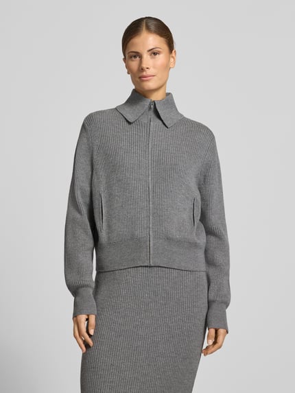 Frauenmodel trägt Regular Fit Strickjacke aus Schurwolle Modell 'FIBIONE' von DRYKORN, graue Farbe, gerippte Textur, Reißverschluss, reine Schurwolle, Nahaufnahme Oberkörper.