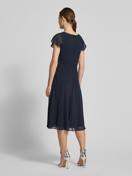 Frauenmodel trägt Knielanges Cocktailkleid mit Ziersteinbesatz von Christian Berg Cocktail in Dunkelblau, Chiffon, taillierter Schnitt, Rückansicht Ganzkörperansicht.