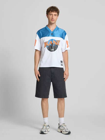 Männermodel trägt Herren T-Shirt Trikot-Shirt mit Logo- und Motiv-Prints Modell 'OG Sport College' von KARL KANI im Boxy-Fit mit V-Ausschnitt, Label-Patch, Weiß und Blau, Rückenlänge 64 cm, Ganzkörperansicht von vorne.