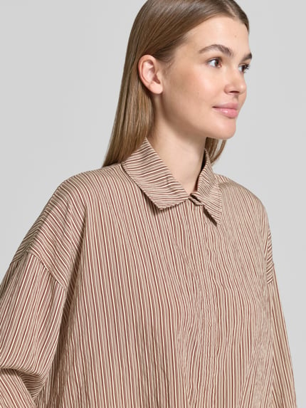 Vrouwenmodel draagt Boxy Fit Bluse uit viscose-mix model 'FIONEL' van DRYKORN met streeppatroon, overhemdkraag, verborgen knooplijst, tunnelkoord, crème en roestkleurige tinten, close-up bovenlichaam.