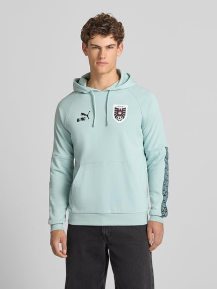 Mężczyzna model nosi Hoodie mit Känguru-Tasche Modell 'ÖFB KING' von PUMA PERFORMANCE, mieszanka bawełny, krój regular fit, długość tyłu w rozmiarze M 70 cm, długość rękawów w rozmiarze M 70 cm, jasnoniebieski, prosty fason, kieszeń kangurka, widok całej sylwetki z przodu.