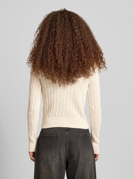 Frauenmodel trägt Strickpullover mit Zopfmuster und Label-Stitching von REVIEW, Baumwoll-Mix, Regular-Fit, Rundhalsausschnitt, Rückenlänge 52 cm, Ärmellänge 69 cm, cremefarben, feine Strickstruktur, dunkle Jeans, Ganzkörperansicht Rückansicht.