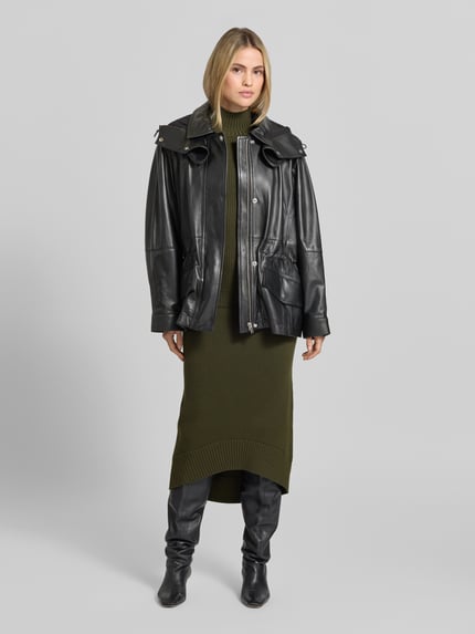 Frauenmodel trägt Funktionsjacke Modell 'GROOVE' von IRO in schwarzer Lederoptik mit metallischen Akzenten und Kapuze, Ganzkörperansicht von vorne.