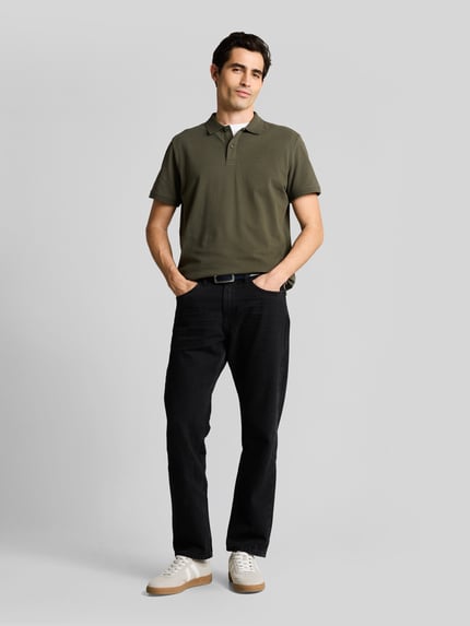 Męski model nosi Regular Fit Poloshirt aus reiner Baumwolle od Tom Tailor, oliwkowa zieleń, krój regular fit, krótki rękaw, pikowana bawełna, kołnierzyk polo, zapięcie na dwa guziki, logo haftowane, gładki wzór, boczne rozcięcia, widok całej sylwetki z przodu.