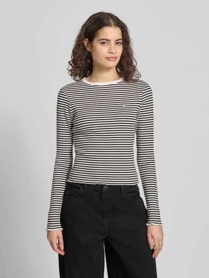 Vrouwenmodel draagt Slim Fit Langarmshirt aus Baumwoll-Mix van Tommy Jeans in zwart-wit gestreept fijnrib, met logo, ronde hals, frill afwerking, frontale totaalopname.