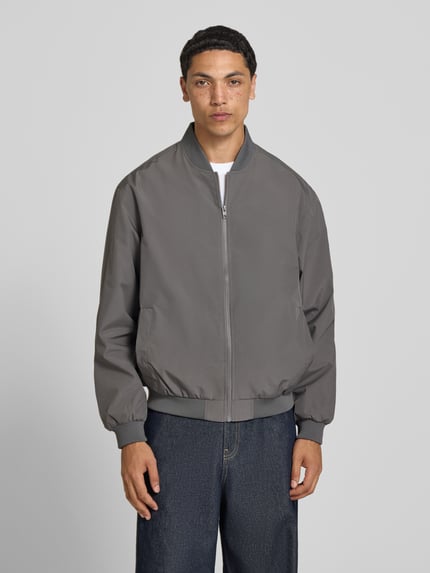 Męski model nosi bomberkę ze stójką model 'CHARGE' od Jack & Jones, krój regular fit, szary, zamek błyskawiczny, długość tyłu 67 cm, szerokość ramion w rozmiarze M 53 cm, szerokość klatki piersiowej w rozmiarze M 62 cm, długość rękawów w rozmiarze M 65 cm, widok całej sylwetki z przodu.