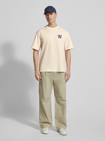 Männermodel trägt Relaxed Fit T-Shirt mit Logo-Print Modell 'Liam' von Wood Wood in Beige, Rundhalsausschnitt, Vorderansicht Ganzkörperansicht.