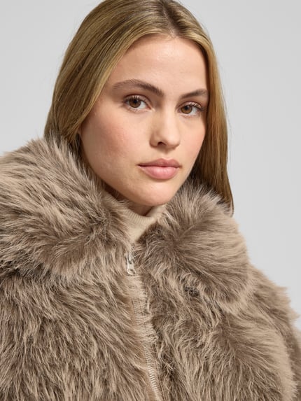 Modelka kobieca nosi Jacke aus Kunstfell mit Reißverschluss od Stefanel w beżowym kolorze, miękka tekstura, zbliżenie na górną część ciała. Regular Fit, kołnierz, zamek, szczegóły: długość 54 cm, szerokość ramion 46 cm, klatka piersiowa 51 cm, rękawy 58 cm.