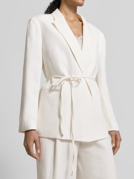 Vrouwenmodel draagt een witte Anzugblazer met reverskraag van Jake*s Collection, taillering, stoffen ceintuur, paspelzakken, close-up bovenlichaam.