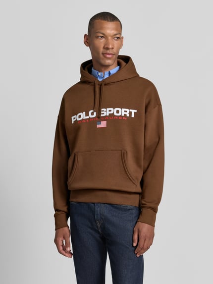 Männermodel trägt Relaxed Fit Hoodie aus Baumwoll-Mix von Polo Ralph Lauren in Braun mit weißen Schriftzügen, Ganzkörperansicht von vorne.
