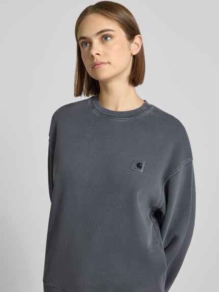 Frauenmodel trägt Relaxed Fit Sweatshirt aus reiner Baumwolle von Carhartt Work In Progress in Dunkelgrau mit Logo-Patch, Nahaufnahme Oberkörper.